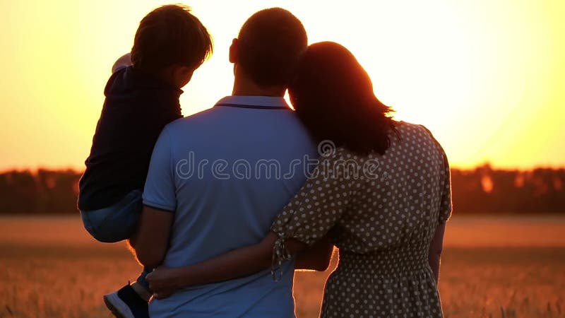 Happy family watching the sunset, standing in a wheat field. A man holding a child in his arms. A woman hugs a man