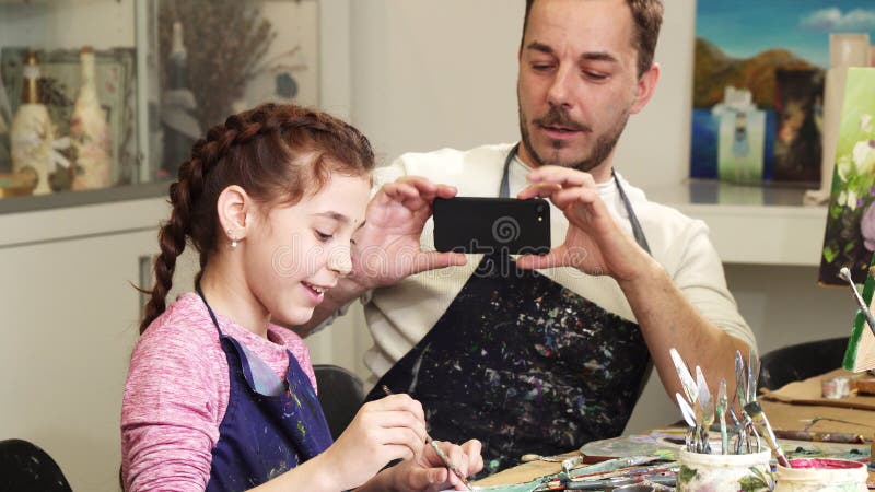 Adorable little girl drawing at art studio her loving happy father smiling cheerfully taking photos of his daughter with a smart phone family technology happiness love bonding artist painting. Adorable little girl drawing at art studio her loving happy father smiling cheerfully taking photos of his daughter with a smart phone family technology happiness love bonding artist painting.