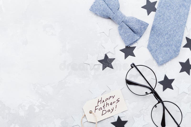 Happy Fathers Day background with greeting tag, glasses, necktie, bowtie and star confetti on table top view. Flat lay composition.