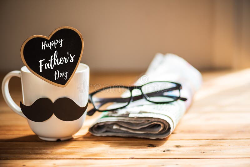 Happy fathers day concept. coffee cup with black paper mustache, heart tag with Happy father`s day text and newspaper, glasses on wooden table background.