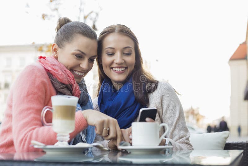 Happy female friends using cell phone at sidewalk cafe