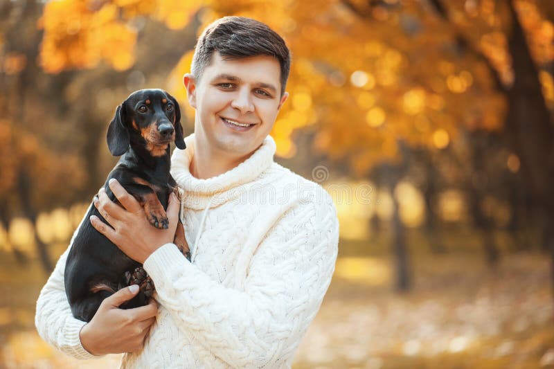 Happy free time with beloved dog! Handsome young man staying in autumn park smiling and holding cute puppy dachshund.