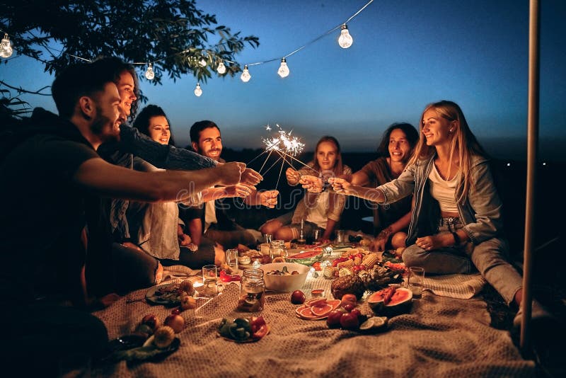 Happy friends having fun with fire sparkles. Young people millennials camping at picnic after sunset. Youth friendship concept on night mood