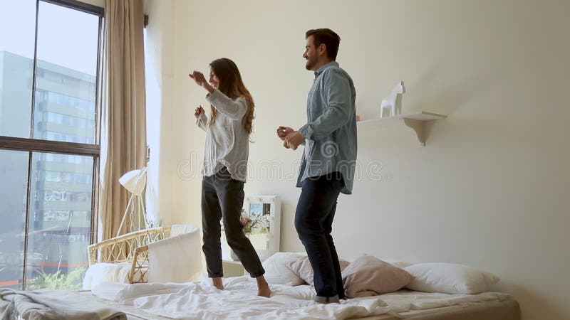 Happy funny young family couple dancing jumping on bed mattress
