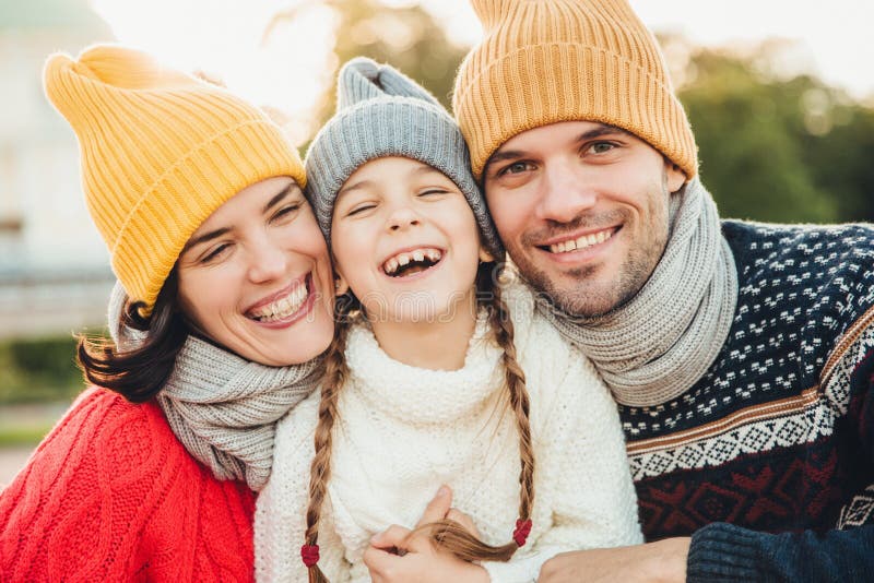 Happy girl with pigtails, wears warm knitted sweater, stands between father and mother, laugh happilly, have sincere smiles on the royalty free stock images Happy girl with pigtails, wears warm knitted sweater, stands between father and mother, laugh happilly, have sincere smiles on the royalty free stock images