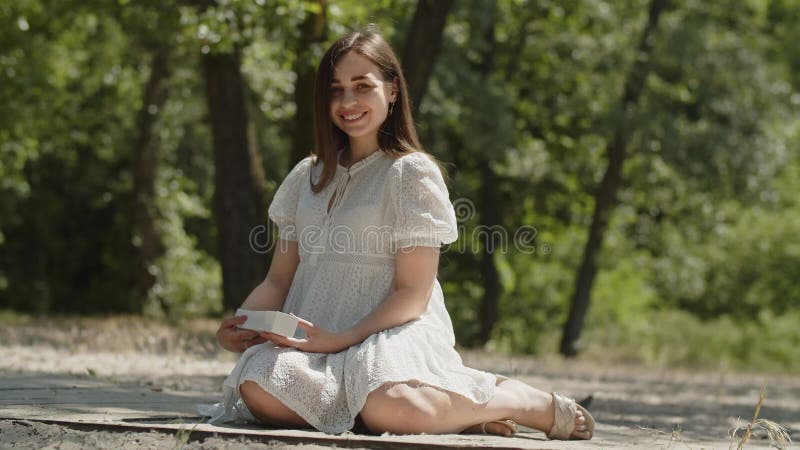 A happy girl in positive mood sits on the sand in a summer forest. A young lady in a white dress holds a box with a