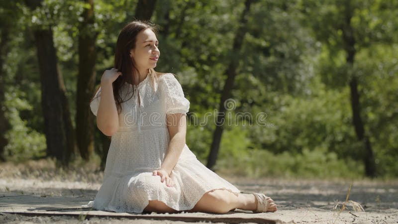 A happy girl in positive mood sits on the sand in a summer forest. A young lady in a white dress holds a box with a