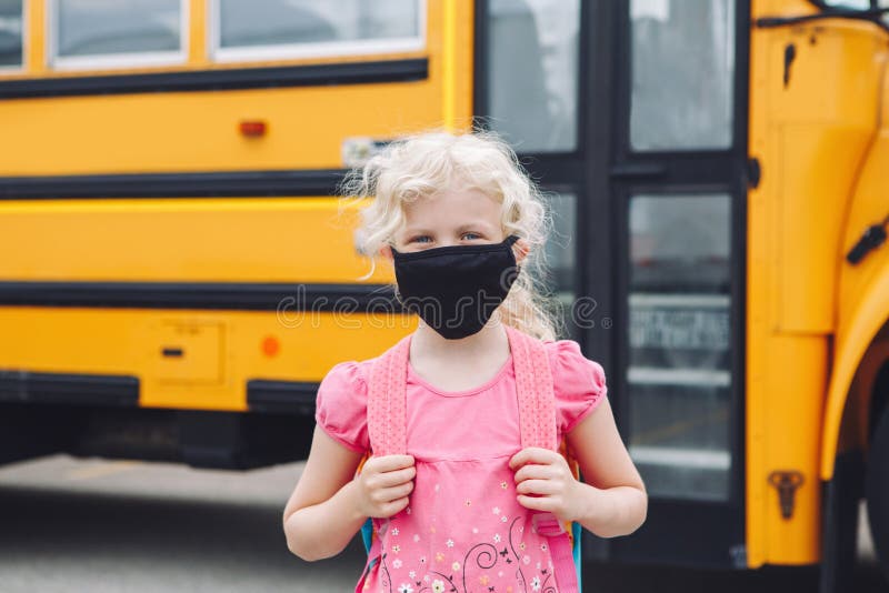 Happy girl student in cloth face mask by yellow school bus outdoor. Kid with personal protective equipment on face. Education and stock photography Happy girl student in cloth face mask by yellow school bus outdoor. Kid with personal protective equipment on face. Education and stock photography