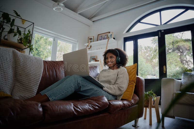 Happy girl wearing headphones watching and listening videos on laptop sitting on couch at home royalty free stock image