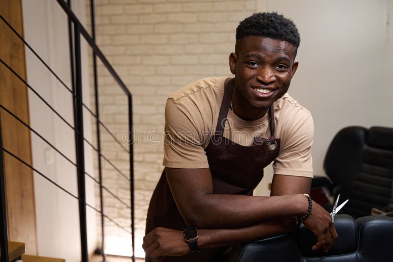 Happy Hairdresser In Working Apron Stands Leaning On Hairdressing Chair Happy Hairdresser In Working Apron Stands Leaning On Hairdressing Chair