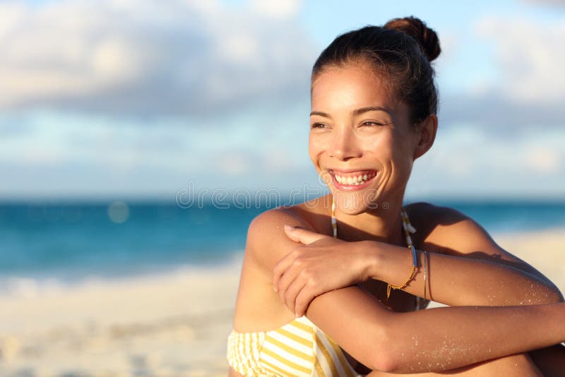 Happy healthy young Asian chinese woman smiling on beach enjoying relax lifestyle sitting in swimwear. Beautiful mixed race tourist in hair bun and golden jewelry at sunset on Caribbean travel. Happy healthy young Asian chinese woman smiling on beach enjoying relax lifestyle sitting in swimwear. Beautiful mixed race tourist in hair bun and golden jewelry at sunset on Caribbean travel.