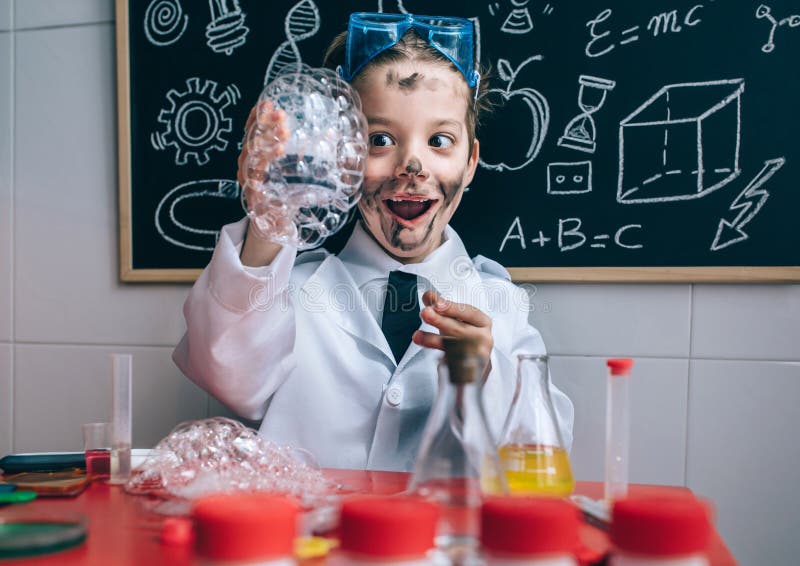 Portrait of excited little boy scientist with dirty face holding glass with soap foam against of blackboard with drawings. Portrait of excited little boy scientist with dirty face holding glass with soap foam against of blackboard with drawings