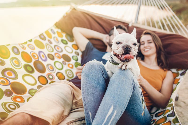 Happy hipster couple with bulldog relaxing in hammock on the beach in sunset light, summer vacation. stylish family with funny dog