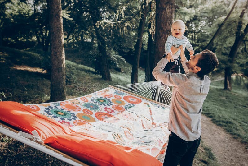 Happy hipster father playing with his cute little son on hammock in summer sunny park. stylish dad with kid relaxing in forest and