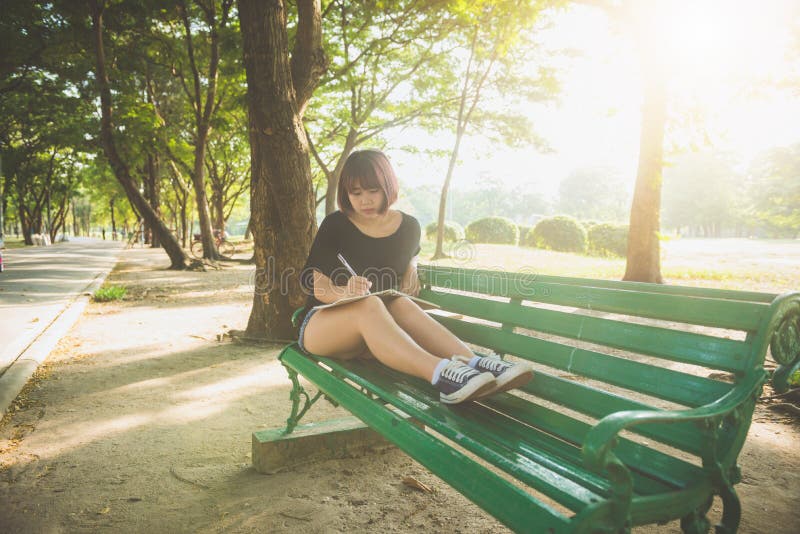 Happy hipster young asian woman writing into her diary in park. Happy hipster young asian woman working on notebook in park.