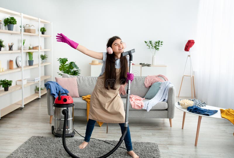 Happy Indian teen girl cleaning her home, wearing headphones, using vacuum cleaner as microphone and having fun