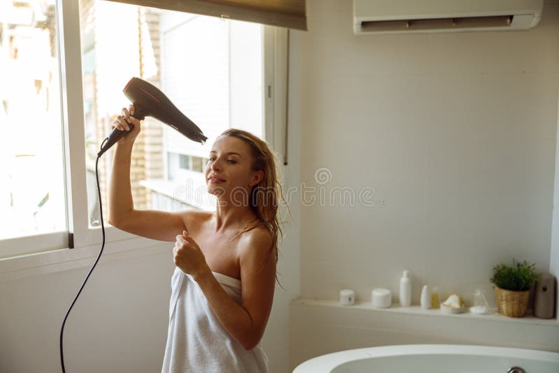 Happy joyful young pretty female in towel drying hair with dryer after taking bath. Relax morning.