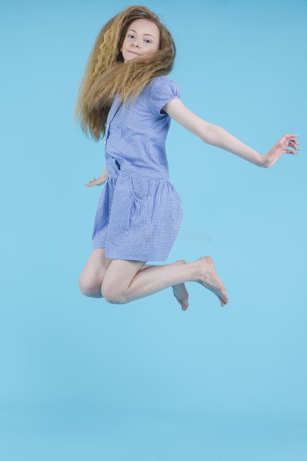 Happy Jumping Teenager Blond Girl in Long Blue Dress Against Blue Background
