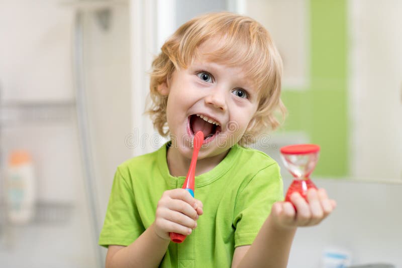 Happy child boy brushing teeth near mirror in bathroom. He is monitoring lasting of cleaning action with hourglass.