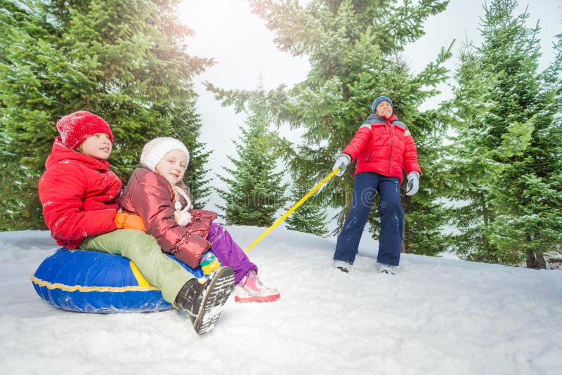 Happy kids sit on snow tube and other pulling them in winter during day in the fir tree forest