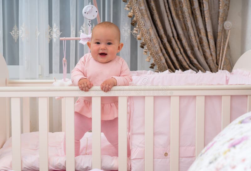 Happy baby standing in crib in pink bedroom scene.