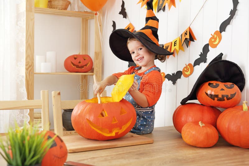 Happy laughing child girl in a witches hat cut a pumpkin for Hal