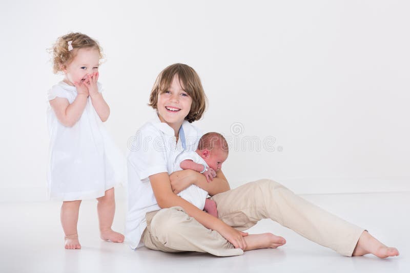 Happy laughing kids, a teen age boy and his adorable curly toddler sister playing with their new born baby brother in a white room with white clothes. Happy laughing kids, a teen age boy and his adorable curly toddler sister playing with their new born baby brother in a white room with white clothes