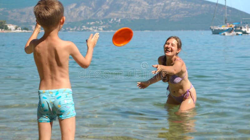 Happy laughing woman playing frisbee with her son on sunny sea beach during summer holidays