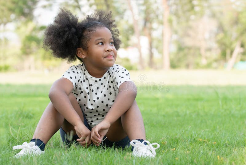 Happy little African child girl smiling and sitting on green grass with afro twin tails hair at park