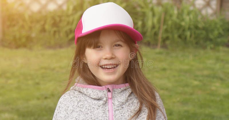 Happy little cute girl in trucker cap smiling and looking at camera while standing in summer garden