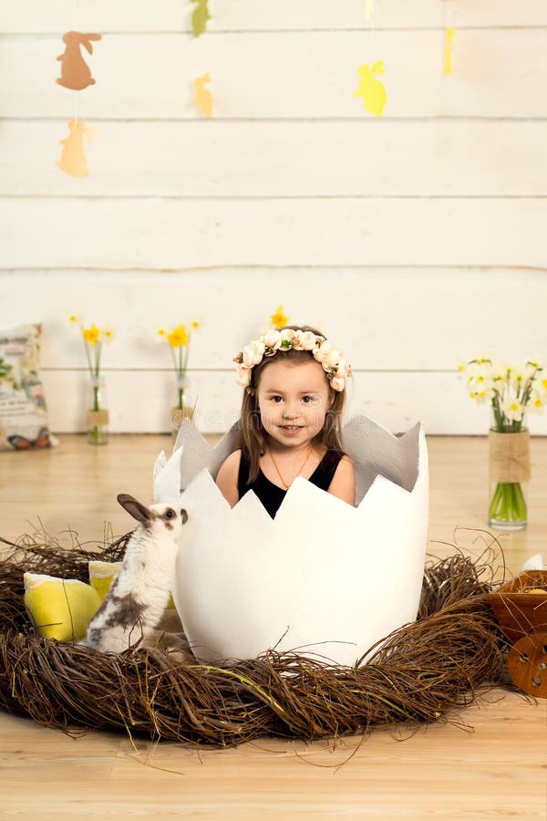 A happy little girl with flowers on her head is sitting in a decorative egg with cute fluffy Easter bunny. Easter egg in the stock photos A happy little girl with flowers on her head is sitting in a decorative egg with cute fluffy Easter bunny. Easter egg in the stock photos