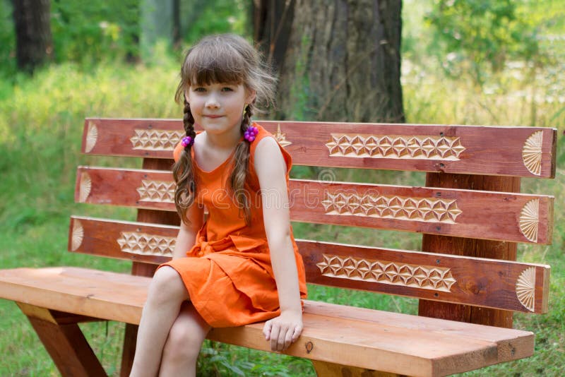 Happy little girl in orange sits on wooden beanch at summer