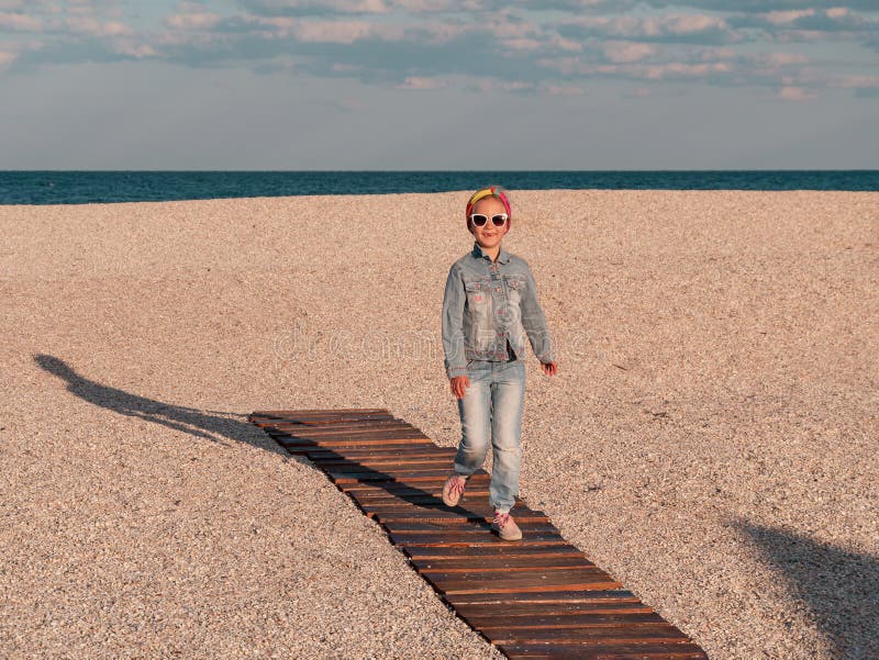 Happy little girl walking alone on wooden boardwalk on seascape background blue sky. Child resting running on sand beach