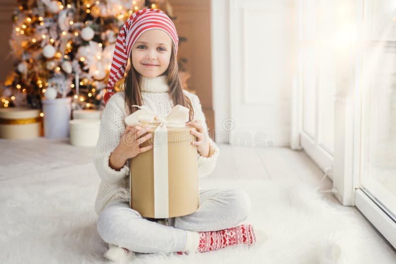 Happy little small child wears white knitted sweater holds gift sits in cozy room against New Year tree, feels comfort, glad to re royalty free stock photo Happy little small child wears white knitted sweater holds gift sits in cozy room against New Year tree, feels comfort, glad to re royalty free stock photo