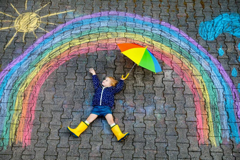 Happy little toddler girl in rubber boots with rainbow sun and clouds with rain painted with colorful chalks on ground