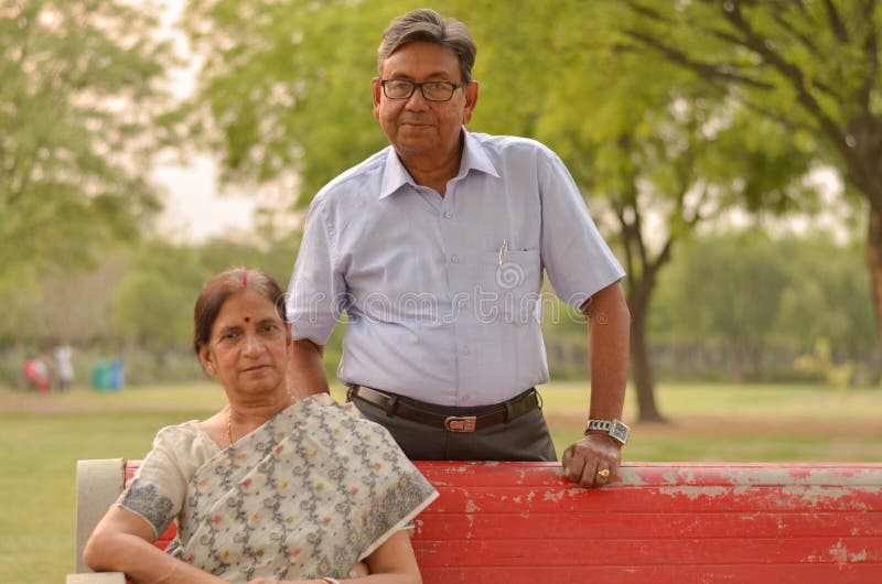 Happy looking retired senior Indian man and woman wearing saree couple smiling sitting on a red park bench in an outdoor setting