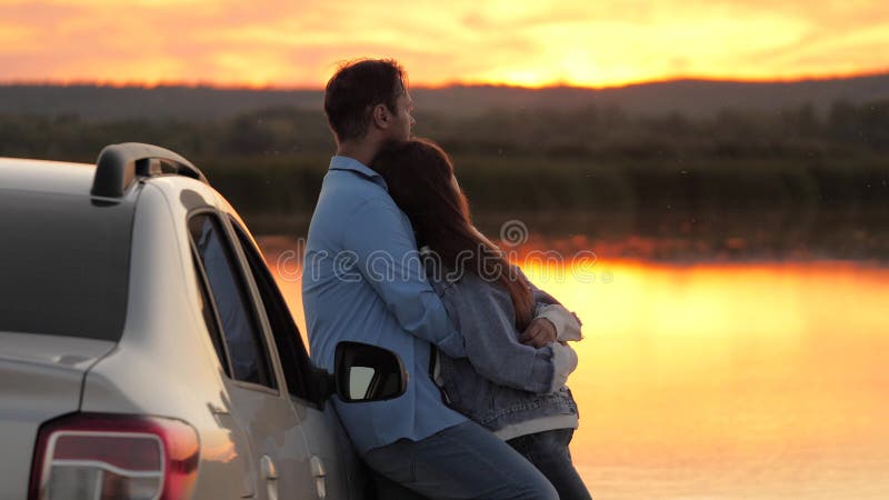 Happy lovers travelers man and woman stand next to car and admire beautiful sunset on beach. Tourists travel by car, hug