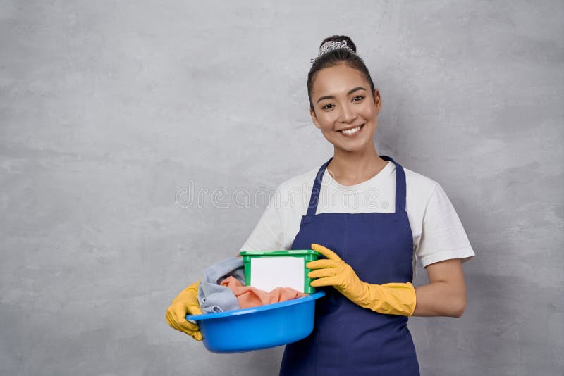 Happy maid woman in uniform holding basket with laundry and green plastic box with washing capsules, smiling at camera royalty free stock image