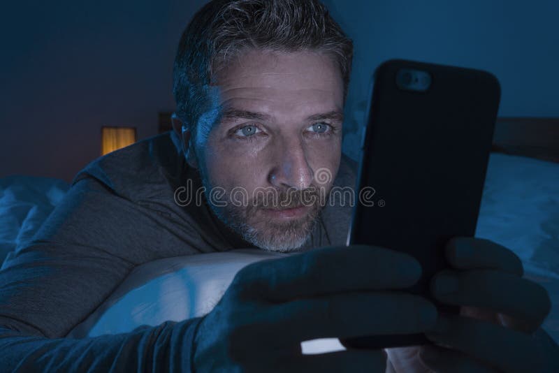 Happy man with blue eyes lying on bed late at night in dark light networking on mobile phone or online dating smiling relaxed
