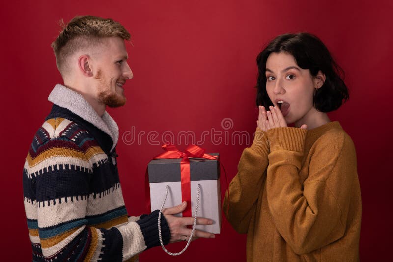Happy man presents Christmas gift box to surprised girlfriend on red background