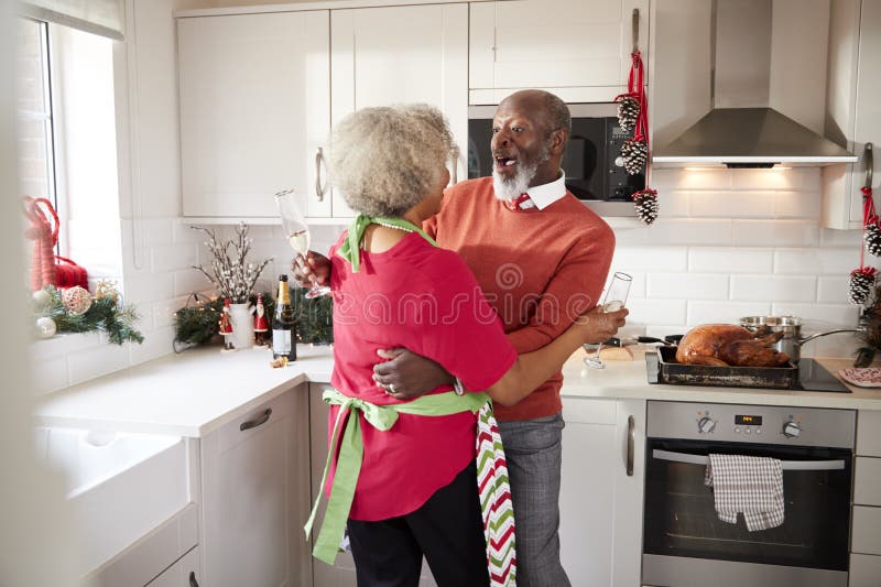 Happy mature black couple holding champagne glasses, laughing and embracing in the kitchen while preparing meal on Christmas morni