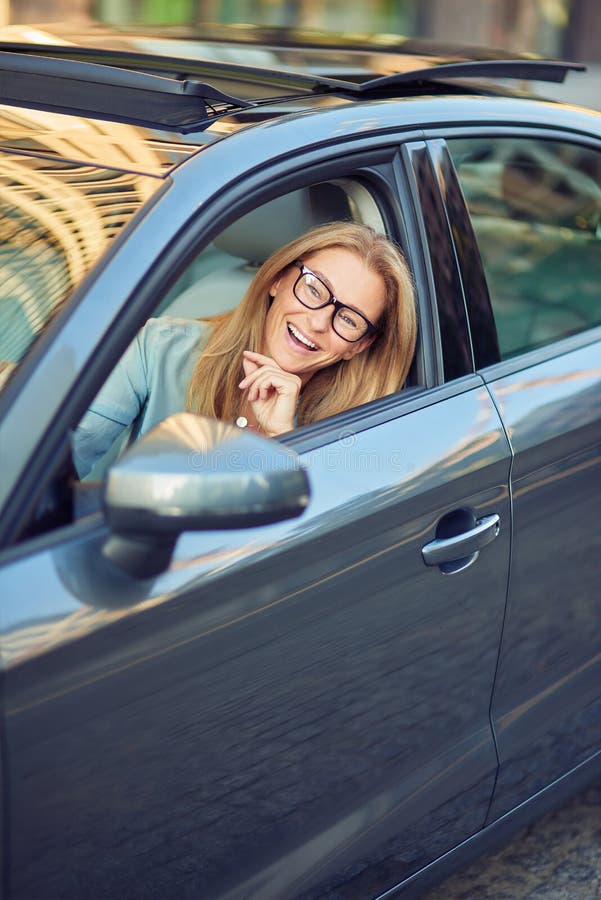 Happy mature business woman wearing eyeglasses driving a car, sitting behind steering wheel, looking through the window