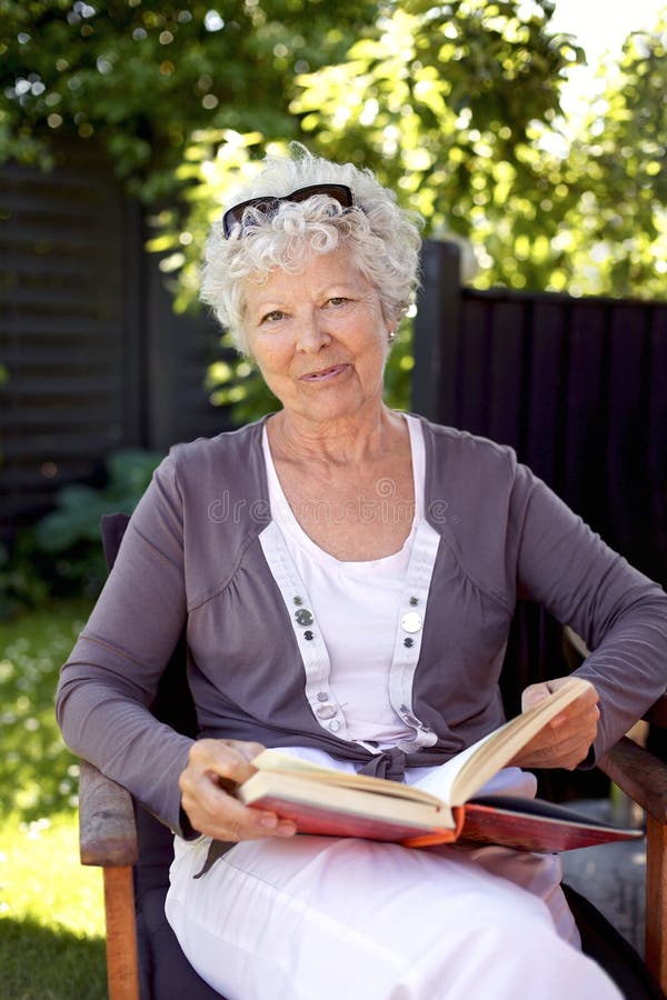 Happy mature woman sitting on a chair in garden with a book looking at camera smiling - Elder woman reading outdoors. Happy mature woman sitting on a chair in garden with a book looking at camera smiling - Elder woman reading outdoors