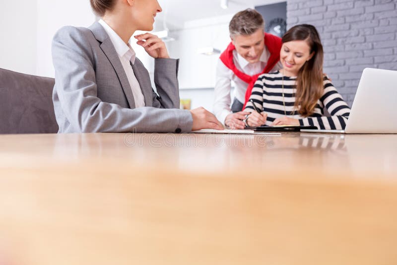Happy man looking at woman signing contract while sitting with female realtor at table in apartment stock photo Happy man looking at woman signing contract while sitting with female realtor at table in apartment stock photo