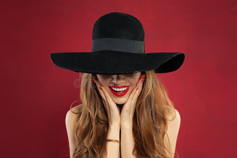 Happy model woman with red lips makeup and manicure on red background. Pretty surprised model in black hat portrait