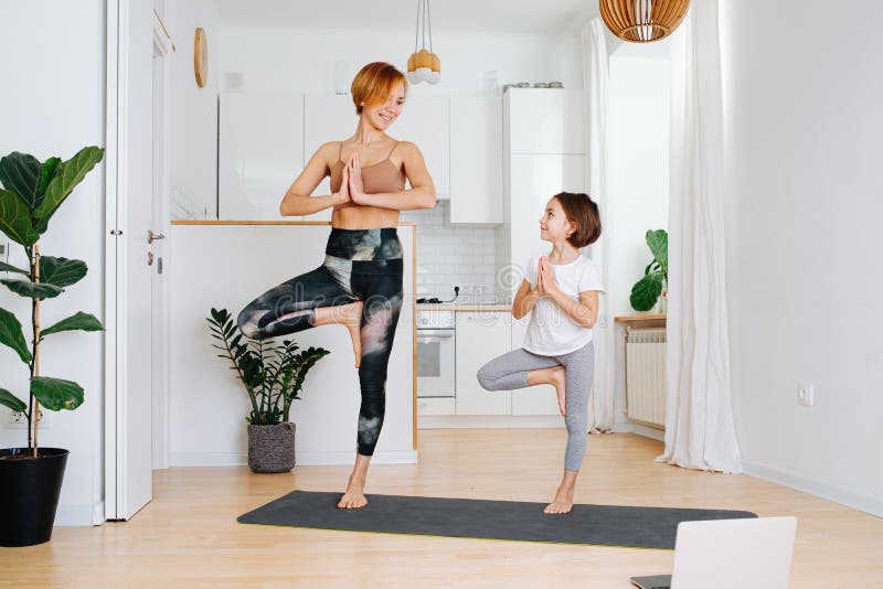 Mom and daughter doing yoga on a yoga mat at home. They both staying onone leg, smiling at each other. Mom and daughter doing yoga on a yoga mat at home. They both staying onone leg, smiling at each other