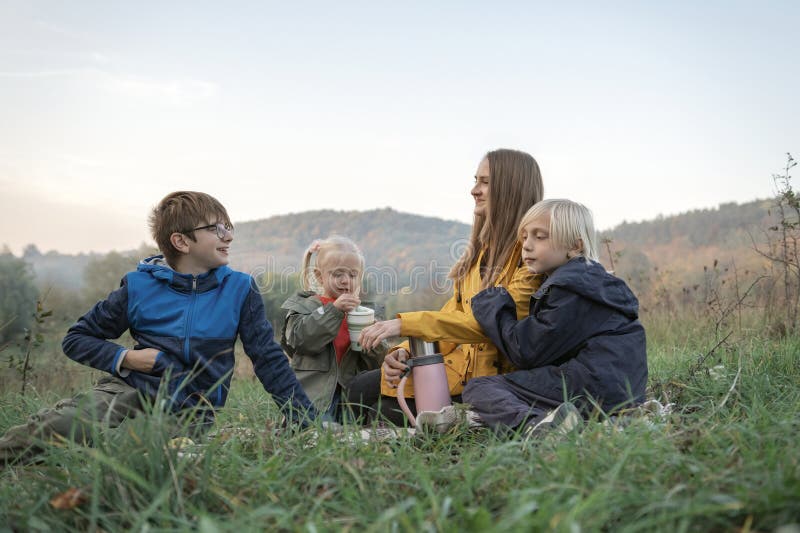 Happy mom and three kids have fun at picnic. Two boys and girl with mother drink hot drink on mountains background