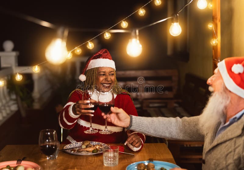 Happy multiracial senior friends toasting with red wine glasses during Christmas holidays dinner celebration on patio house