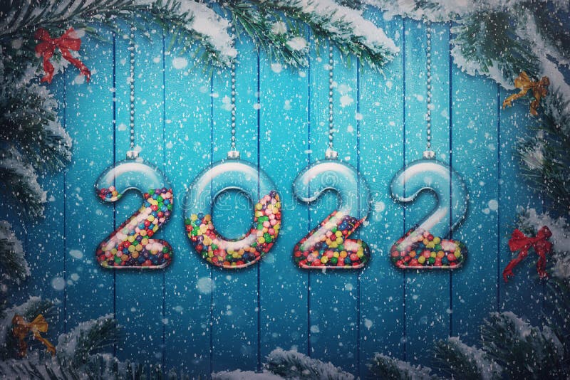 Happy New Year 2022 holiday background. Set of transparent numbers made of glass filled with multi colored candy and sweets hang. On snowy Christmas tree royalty free stock photo