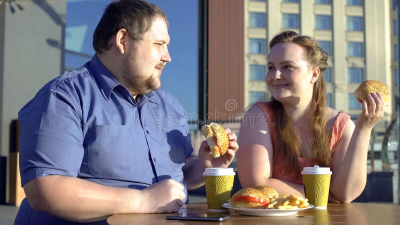Happy obese couple eating hamburgers on romantic date, flirting in outdoor cafe royalty free stock photo Happy obese couple eating hamburgers on romantic date, flirting in outdoor cafe royalty free stock photo